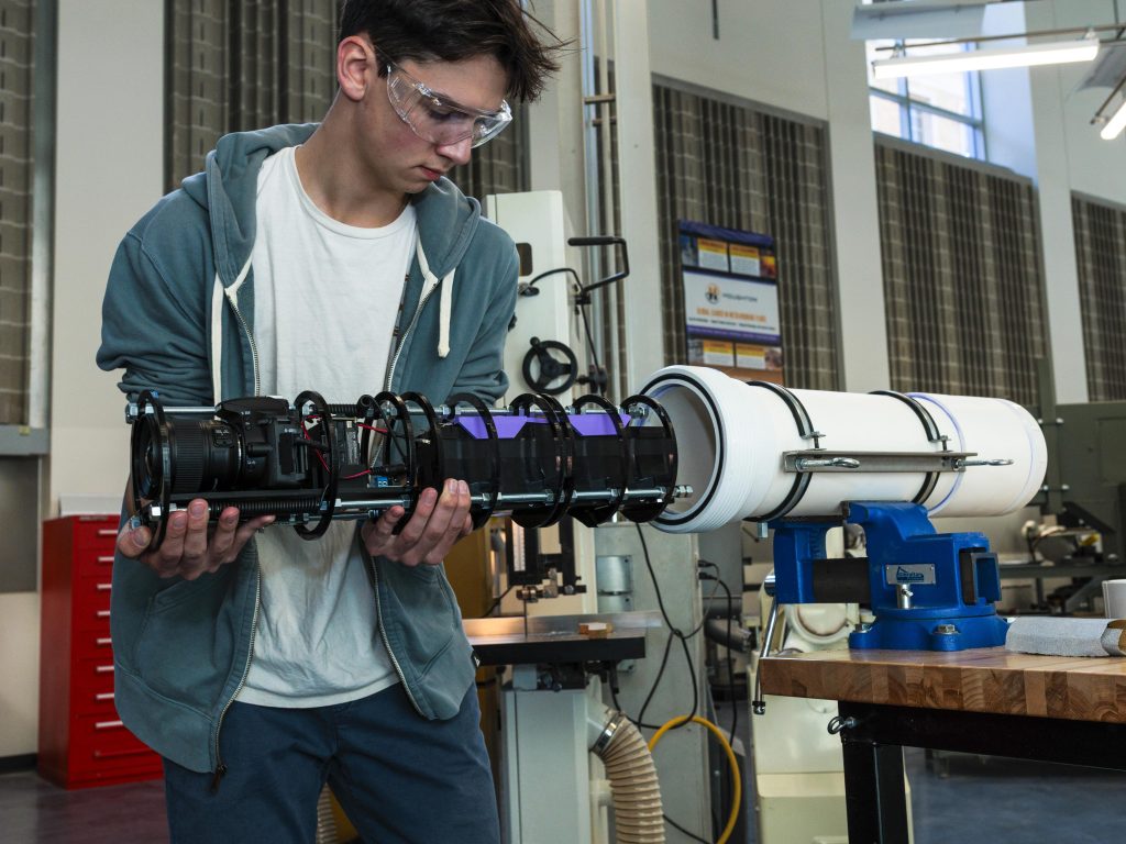 Soren Goldsmith in safety goggles holds a custom-built camera designed for underwater photography, showcasing engineering and technology in a workshop setting.